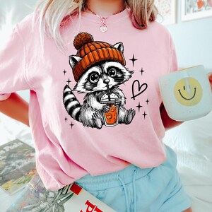 K&ouml;nnte beinhalten: Ein pinkfarbenes T-Shirt mit einem Cartoon-Waschb&auml;ren, der eine orangefarbene M&uuml;tze tr&auml;gt und eine Tasse Kaffee h&auml;lt. Der Waschb&auml;r ist von Sternen und einem Herzen umgeben.