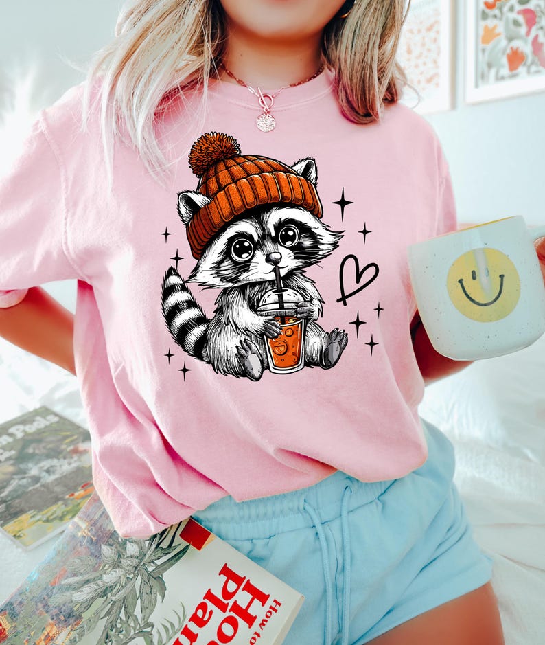 K&ouml;nnte beinhalten: Ein pinkfarbenes T-Shirt mit einem Cartoon-Waschb&auml;ren, der eine orangefarbene M&uuml;tze tr&auml;gt und eine Tasse Kaffee h&auml;lt. Der Waschb&auml;r ist von Sternen und einem Herzen umgeben.