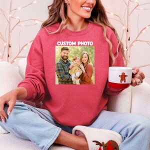 Puede incluir: Una sudadera roja con una foto familiar y el texto "CUSTOM PHOTO". La persona sostiene una taza blanca y roja con un dise&ntilde;o de hombre de jengibre. La persona lleva vaqueros azules y zapatillas de reno.