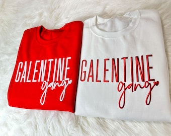 May include: Two red and white sweatshirts with the text "Galentine gang" embroidered on them. The red sweatshirt has white text and the white sweatshirt has red text.