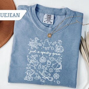 May include: Bluejean colored t-shirt with a crew neck and short sleeves. The shirt features a white graphic of Halloween-themed geese and the phrase "just a spooky goose." A gold necklace is draped across the shirt.
