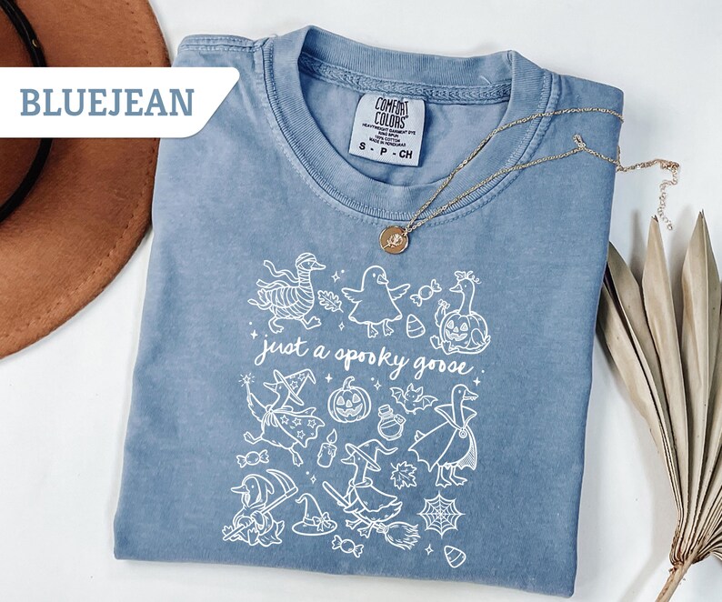 May include: Bluejean colored t-shirt with a crew neck and short sleeves. The shirt features a white graphic of Halloween-themed geese and the phrase "just a spooky goose." A gold necklace is draped across the shirt.