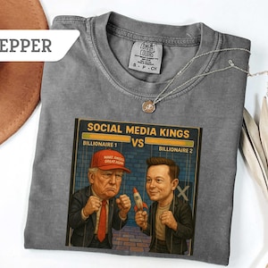 Puede incluir: Una camiseta gris Comfort Colors con un gráfico de dos hombres, etiquetados como "Billionaire 1" y "Billionaire 2", con el texto "Social Media Kings". Un hombre lleva una gorra roja "Make America Great Again". Una cadena de plata está sobre la camiseta.