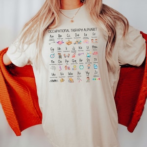 May include: A white t-shirt with a graphic of an occupational therapy alphabet. The alphabet is illustrated with icons representing different aspects of occupational therapy, such as adaptive equipment, brain gym, coordination, development, executive functioning, fine motor, goals, helpful, independence, just right challenge, key group, leisure, memory, neurodiversity,  orthodontics, play, quality of life, routine, self-feeding, toileting, universal design, visual motor, wheelchair, exercise, yoga, and zones of regulation.