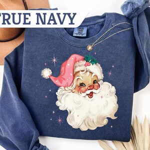 May include: Navy blue sweatshirt featuring a vintage Santa Claus graphic. The Santa Claus illustration has a pink hat, white beard, and rosy cheeks. The text "TRUE NAVY" is displayed across the top of the image. A gold necklace is draped across the sweatshirt.