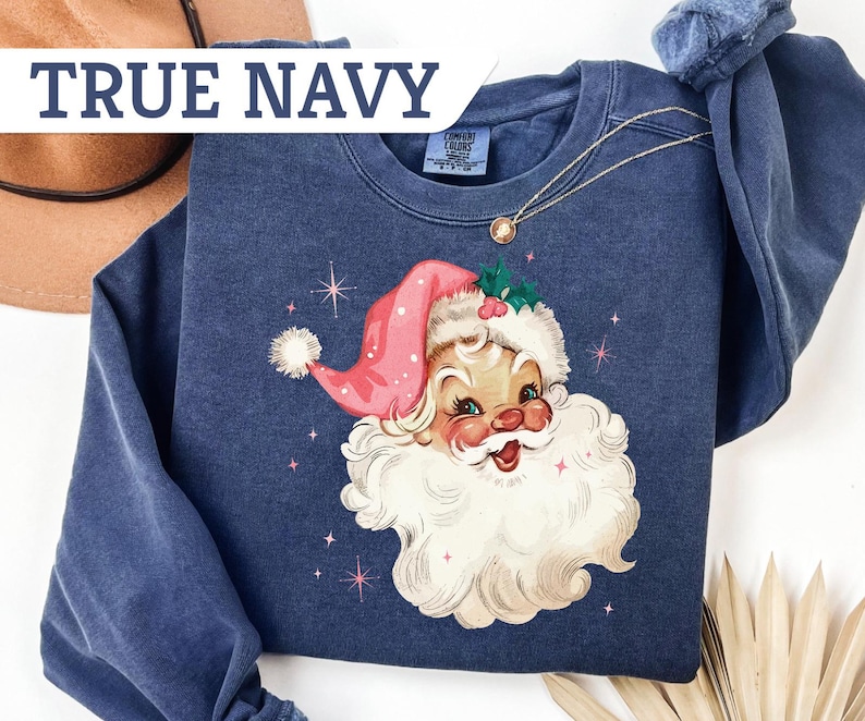 May include: Navy blue sweatshirt featuring a vintage Santa Claus graphic. The Santa Claus illustration has a pink hat, white beard, and rosy cheeks. The text "TRUE NAVY" is displayed across the top of the image. A gold necklace is draped across the sweatshirt.