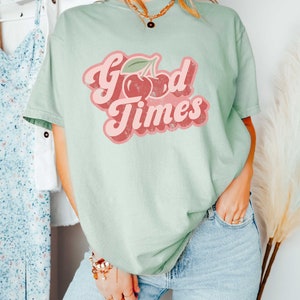 Pu&ograve; includere: T-shirt verde chiaro con la scritta "Good Times" in un font retr&ograve;, con una grafica di ciliegie. La maglietta &egrave; abbinata a jeans azzurri. Lo stile generale &egrave; casual e rilassato.