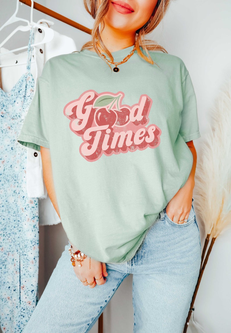Pu&ograve; includere: T-shirt verde chiaro con la scritta "Good Times" in un font retr&ograve;, con una grafica di ciliegie. La maglietta &egrave; abbinata a jeans azzurri. Lo stile generale &egrave; casual e rilassato.