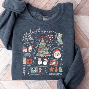 May include: A dark blue sweatshirt with a festive Christmas graphic. The design includes a decorated tree, candy canes, stockings, and the phrase "tis the season." Other elements include Christmas lights, Santa, and baking cookies.
