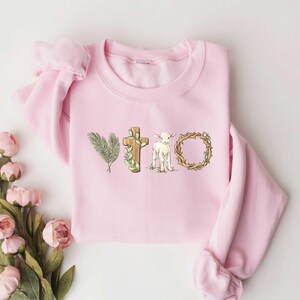 May include: Pink sweatshirt with a graphic design of a cross, a lamb, and a crown of thorns. The graphic is in shades of brown, green, and white.
