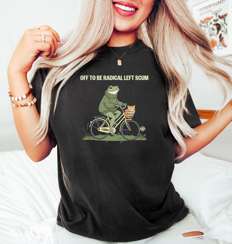 May include: Black t-shirt with the text "OFF TO BE RADICAL LEFT SCUM." The shirt features a cartoon frog riding a bicycle with a cat in a basket. The frog is wearing a green sweater. The bicycle is yellow.