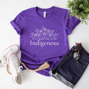 May include: Purple t-shirt with white text that reads "It's a good day to be Indigenous" and a floral design above the text.