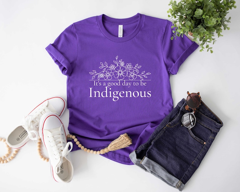 May include: Purple t-shirt with white text that reads "It's a good day to be Indigenous" and a floral design above the text.