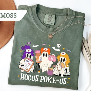 May include: Moss green t-shirt with a Halloween-themed graphic. The design features three ghost nurses with medical supplies and the text "Hocus Poke-Us". A gold necklace is draped across the shirt.