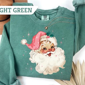 May include: Light green sweatshirt featuring a vintage Santa Claus graphic. The Santa Claus illustration has a pink hat, white beard, and rosy cheeks. The text "LIGHT GREEN" is in a white box in the upper left corner. A gold necklace is visible.