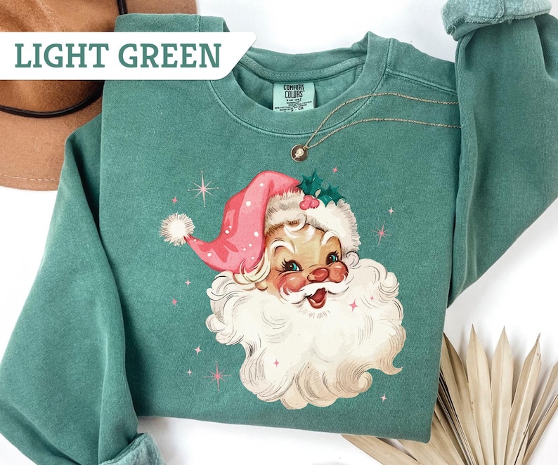 May include: Light green sweatshirt featuring a vintage Santa Claus graphic. The Santa Claus illustration has a pink hat, white beard, and rosy cheeks. The text "LIGHT GREEN" is in a white box in the upper left corner. A gold necklace is visible.