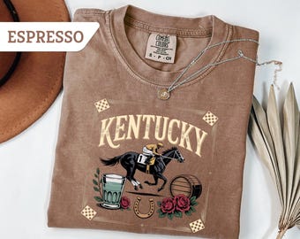 May include: Espresso-coloured t-shirt featuring a horse race graphic, the word "Kentucky", a mint julep, roses, and a horseshoe. The shirt has a crew neck and short sleeves. A silver necklace is draped across the shirt.