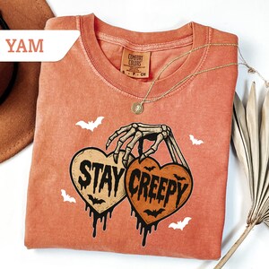 May include: A coral-colored t-shirt with a crew neck and short sleeves. The shirt features a graphic of two heart-shaped pumpkins held by a skeleton hand, with the words "STAY CREEPY" and bat illustrations. A gold necklace is draped across the shirt.