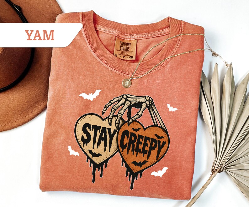 May include: A coral-colored t-shirt with a crew neck and short sleeves. The shirt features a graphic of two heart-shaped pumpkins held by a skeleton hand, with the words "STAY CREEPY" and bat illustrations. A gold necklace is draped across the shirt.
