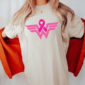 May include: A white t-shirt with a pink ribbon design in the shape of the Wonder Woman logo. The ribbon is a symbol of breast cancer awareness.