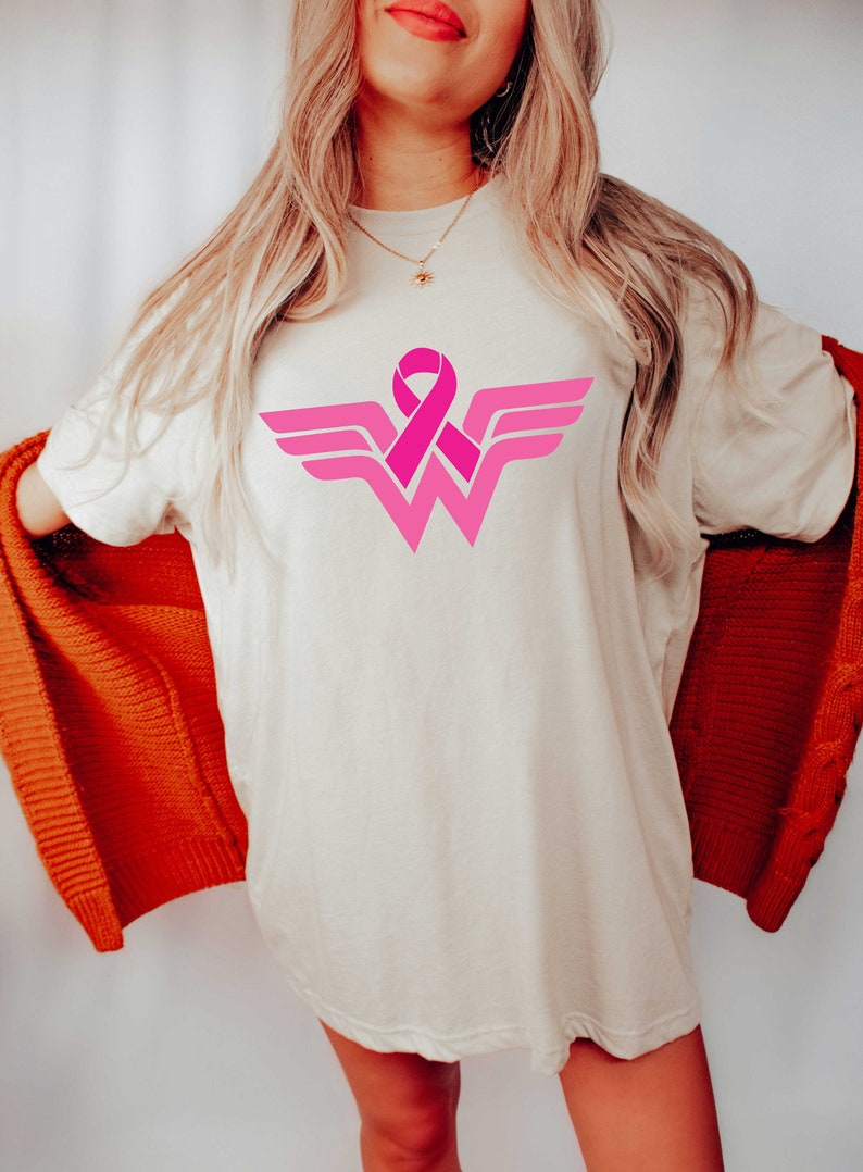 May include: A white t-shirt with a pink ribbon design in the shape of the Wonder Woman logo. The ribbon is a symbol of breast cancer awareness.