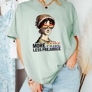 May include: A light green t-shirt featuring a graphic of a woman wearing heart-shaped sunglasses with rainbow colours. The text "More Pride Less Prejudice" is printed below the graphic.