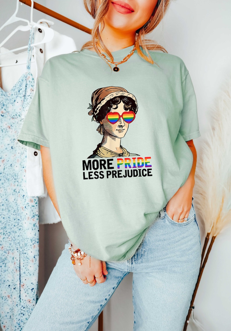 May include: A light green t-shirt featuring a graphic of a woman wearing heart-shaped sunglasses with rainbow colours. The text "More Pride Less Prejudice" is printed below the graphic.