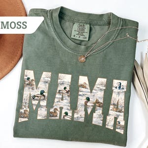May include: A moss green t-shirt with the word "MAMA" printed across the front in a white font. The letters are filled with a duck illustration. A gold necklace and a brown hat are also visible.