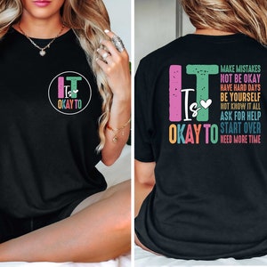 Pode incluir: Camiseta preta com um gr&aacute;fico colorido na frente que diz "It Is Okay To" e um gr&aacute;fico menor nas costas que diz "It Is Okay To" com uma lista de frases incluindo "Make Mistakes", "Not Be Okay", "Have Hard Days", "Be Yourself", "Not Know It All", "Ask For Help", "Start Over" e "Need More Time".