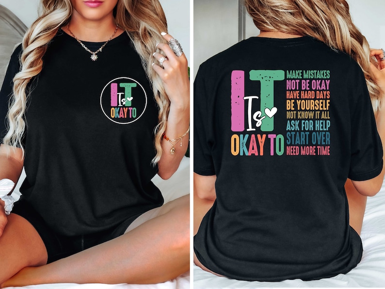 Pode incluir: Camiseta preta com um gr&aacute;fico colorido na frente que diz "It Is Okay To" e um gr&aacute;fico menor nas costas que diz "It Is Okay To" com uma lista de frases incluindo "Make Mistakes", "Not Be Okay", "Have Hard Days", "Be Yourself", "Not Know It All", "Ask For Help", "Start Over" e "Need More Time".