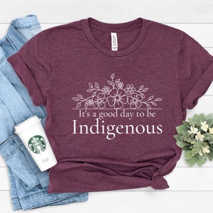 May include: A maroon t-shirt with a white floral design and the text "It's a good day to be Indigenous".