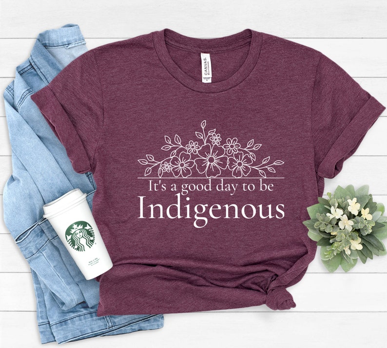 May include: A maroon t-shirt with a white floral design and the text "It's a good day to be Indigenous".