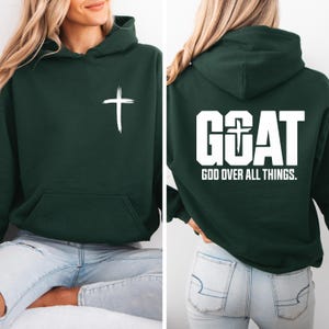 Pode incluir: Moletom verde escuro com uma cruz branca na frente e o texto "GOAT GOD OVER ALL THINGS" nas costas. O moletom tem um bolso frontal e um capuz. O texto e a cruz est&atilde;o em negrito branco.