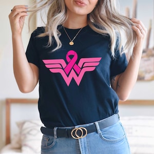 May include: Black t-shirt with a pink ribbon and a pink and white Wonder Woman logo. The logo is a stylized 'W' with wings.