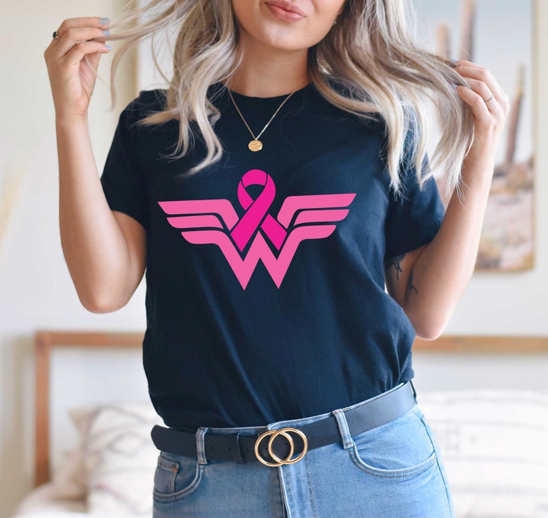 May include: Black t-shirt with a pink ribbon and a pink and white Wonder Woman logo. The logo is a stylized 'W' with wings.