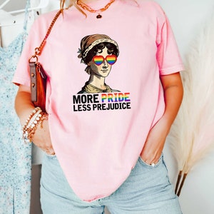 May include: A pink t-shirt featuring a graphic of a woman wearing rainbow heart-shaped sunglasses and the text "More Pride Less Prejudice".