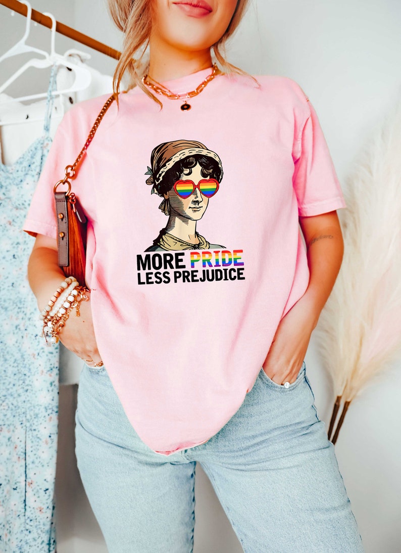 May include: A pink t-shirt featuring a graphic of a woman wearing rainbow heart-shaped sunglasses and the text "More Pride Less Prejudice".