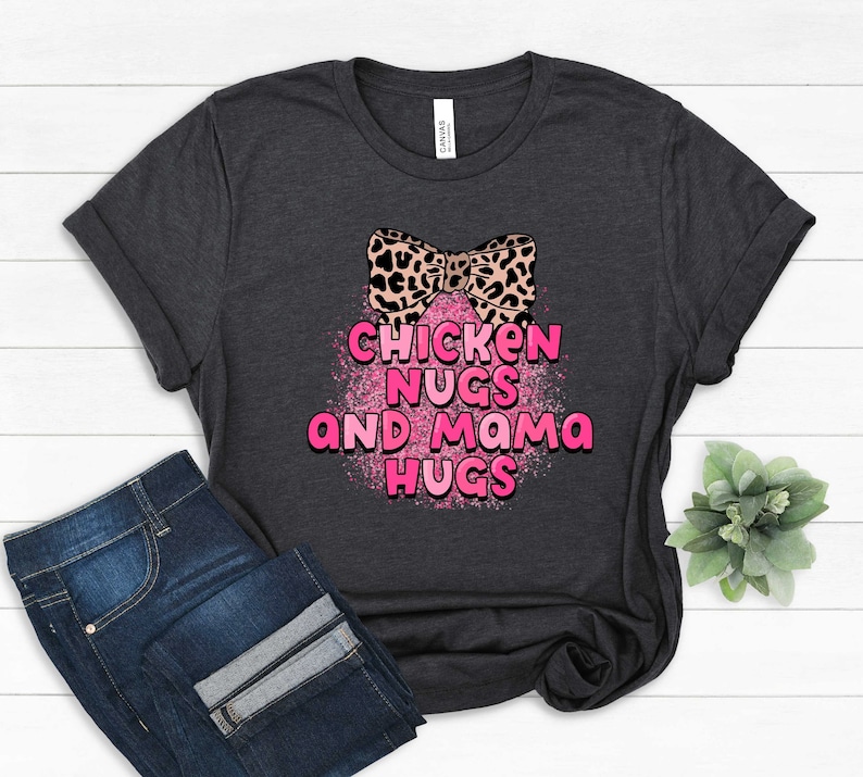 May include: A dark grey t-shirt with a pink and black leopard print bow and the text "Chicken Nugs and Mama Hugs" in pink letters with a pink glitter background.