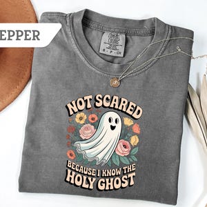 May include: Pepper-colored t-shirt with the text "NOT SCARED BECAUSE I KNOW THE HOLY GHOST" and a cartoon ghost illustration surrounded by flowers. The shirt has a crew neck and short sleeves.