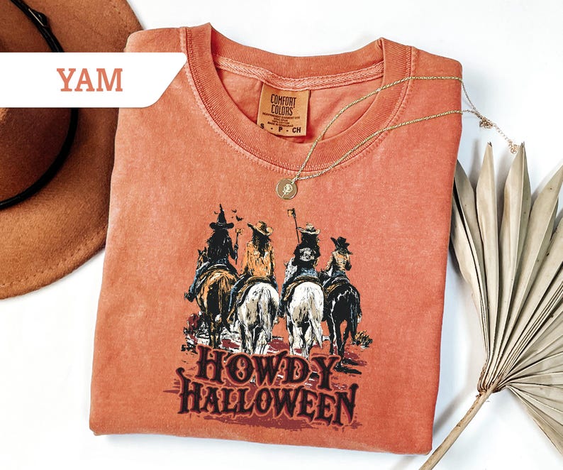 May include: A coral-coloured t-shirt with a graphic of four figures riding horses, and the text "HOWDY HALLOWEEN" in a Western-style font. The shirt has a crew neck and short sleeves. A gold necklace with a pendant is draped on the shirt.