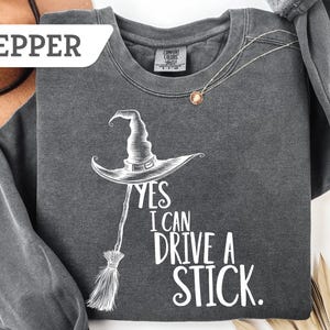 May include: Pepper-colored long-sleeve sweatshirt with a witch hat and broom graphic. The text reads "YES I CAN DRIVE A STICK." A silver necklace with a pendant is draped on the shirt. The sweatshirt is made of a soft material.