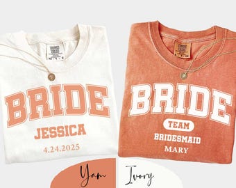 May include: Two folded t-shirts, one ivory and one yam-colored, with the word "BRIDE" in a retro font. The ivory shirt has "JESSICA" and "4.24.2025" printed below. The yam shirt says "TEAM BRIDESMAID MARY". Each shirt has a gold necklace.