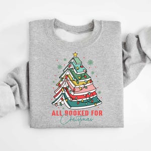 May include: A light gray sweatshirt featuring a colorful Christmas tree design made of books, with a star on top and string lights. The text "ALL BOOKED FOR Christmas" is printed below the tree in red and teal script.