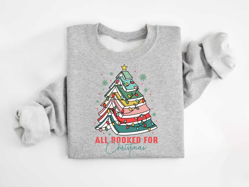 May include: A light gray sweatshirt featuring a colorful Christmas tree design made of books, with a star on top and string lights. The text "ALL BOOKED FOR Christmas" is printed below the tree in red and teal script.