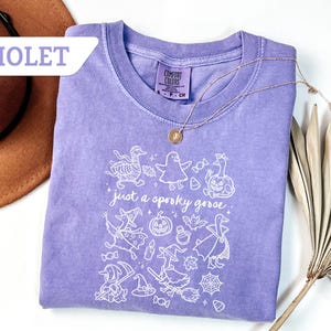 May include: Violet Comfort Colors t-shirt with a white graphic of Halloween-themed geese, witches, and pumpkins. The text "just a spooky goose" is printed below the graphic. A gold necklace is draped on the shirt.