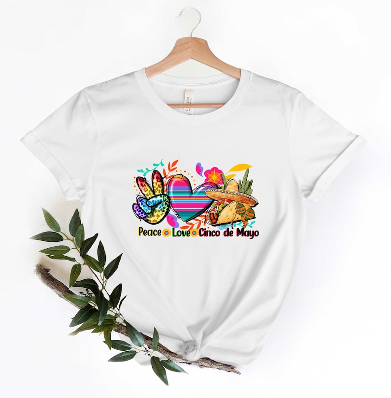 Puede incluir: Camiseta blanca con un dise&ntilde;o colorido de signo de paz, coraz&oacute;n y sombrero. El texto "Peace Love Cinco de Mayo" est&aacute; impreso debajo del dise&ntilde;o.