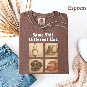 Peut inclure: T-shirt couleur expresso avec le texte "Same Shit. Different Hat." et des illustrations de diff&eacute;rents chapeaux. Un collier argent&eacute; et des baskets blanches sont &eacute;galement visibles. Le t-shirt est pr&eacute;sent&eacute; avec un jean bleu et une plante s&eacute;ch&eacute;e.