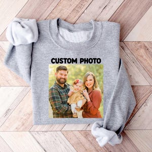 Puede incluir: Una sudadera gris con una foto familiar y el texto "CUSTOM PHOTO" impreso en la parte delantera. La foto muestra a un hombre, una mujer y un beb&eacute;. La sudadera est&aacute; hecha de un material suave y tiene cuello redondo.