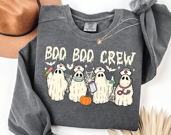 May include: Dark gray crewneck sweatshirt with the text "BOO BOO CREW" and illustrations of ghost characters dressed as nurses. The ghosts are holding medical tools and a small orange pumpkin is at the bottom.