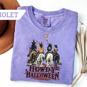 May include: Violet Comfort Colors t-shirt with a graphic of four cowgirls on horseback, and the text "HOWDY HALLOWEEN". A gold necklace and a brown hat are also visible. The t-shirt is a relaxed fit.